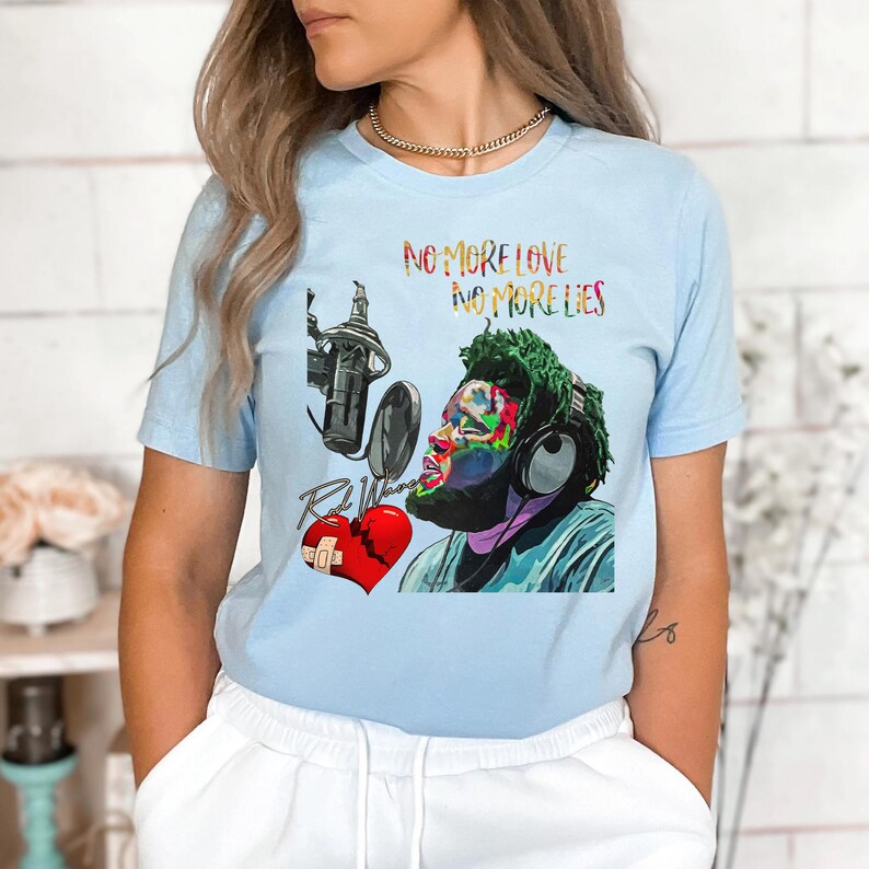 May include: Light blue t-shirt featuring a colorful graphic of a musician with headphones, a microphone, and a broken heart. The text "NO MORE LOVE NO MORE LIES" is printed above the graphic. The artist's name is printed below the heart.