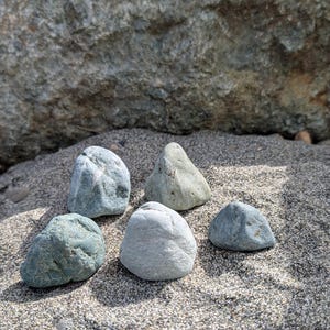 May include: Five smooth, rounded stones of varying shades of gray, blue-green, and pale white are arranged on a textured, dark sandy surface. The stones have natural, irregular shapes and matte textures, contrasting with the coarse sand grains.