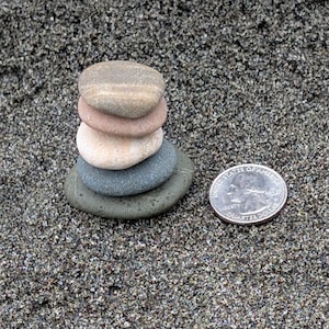 May include: A stack of five smooth, rounded river stones in shades of tan, pink, and gray, arranged vertically. Next to the stone stack is a silver United States quarter coin, which serves as a scale reference for the size of the stones.
