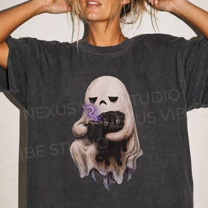 Könnte beinhalten: Ein anthrazitfarbenes Kurzarm-T-Shirt mit einem Grafikdruck eines weißen Geistes, der eine schwarze Tasse mit aufsteigendem violettem Dampf hält und dabei einen kleinen schwarzen Mops im Arm hält. Das Motiv ist mittig platziert.