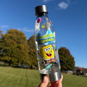May include: A clear plastic water bottle with a silver metal cap and base. The bottle features a colourful graphic of SpongeBob SquarePants, the text SpongeBob SquarePants, and various small character icons printed on the transparent surface.