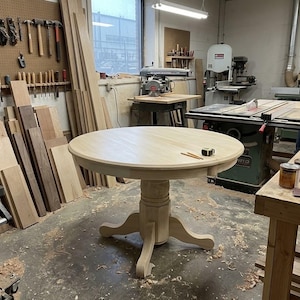 May include: A round, light-colored wooden pedestal table with a smooth, circular top and a central column base featuring four curved feet. Two pencils and a small measuring tape rest on the surface of the unfinished wood table in a workshop.