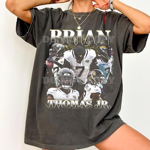 Pode incluir: Uma t-shirt cinzento-carvão de manga curta com uma impressão gráfica de um jogador de futebol americano com camisola branca e o número 7. O texto BRIAN e THOMAS JR. está impresso numa fonte prateada metálica acima e abaixo.
