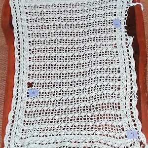 May include: A light green, open-weave crochet blanket with a scalloped edge. The blanket is rectangular and has a delicate, lacy pattern. The blanket is displayed on a brown wooden surface.