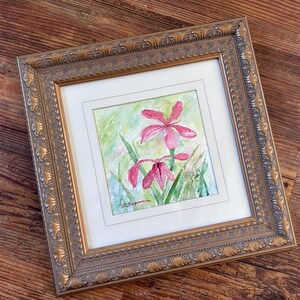 May include: A framed watercolor painting of pink flowers with green foliage. The artwork is set within a white mat and a decorative gold-colored frame. The painting is on a wooden surface.