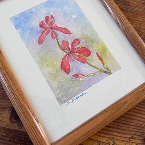May include: Watercolor painting of red flowers with green stems and buds, set against a soft blue and green background. The artwork is framed in a natural wood frame and signed "S. Dupnas" in blue ink.