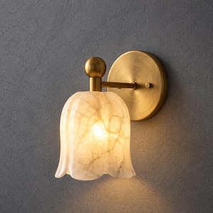 May include: A wall-mounted light fixture featuring a brushed brass circular backplate and a horizontal arm ending in a spherical finial. The lamp shade is shaped like a bell-shaped flower with a white and grey marbled stone-like texture.
