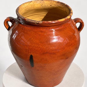 Op de afbeelding: Een grote, ronde, geglazuurde aardenwerken pot met een diepe amberbruine afwerking en een wijde opening. De pot heeft twee kleine, gebogen handvatten aan de zijkanten en een opvallende zwarte verticale markering op de buitenkant, met zichtbare slijtage aan de rand.