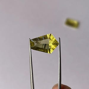 Könnte beinhalten: Ein facettierter, schildförmiger zitronengelber Edelstein, der mit einer Metallpinzette gehalten wird. Der Stein hat ein klares, transparentes Aussehen mit scharfen geometrischen Schliffen, die das Licht reflektieren und eine helle Farbe erzeugen.