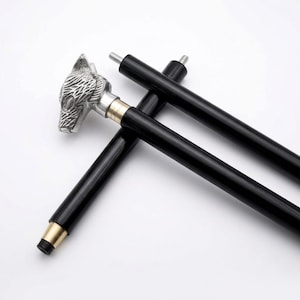 Op de afbeelding: Een wandelstok met een zilverkleurige handgreep in de vorm van een wolvenkop met gedetailleerde vachtstructuur, een zwarte schacht en een messingkleurige kraag. De greep is bevestigd met een metalen bout en heeft een zwarte rubberen dop.