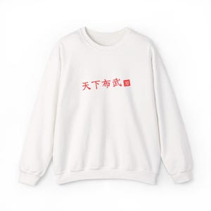 Peut inclure: Un sweat-shirt blanc à manches longues et col rond, orné de caractères kanji japonais rouges imprimés au centre de la poitrine. Le texte indique "天下布武", suivi d'un petit carré rouge contenant un seul caractère kanji blanc.