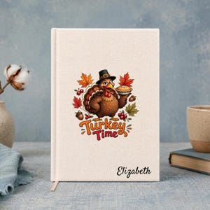 Peut inclure: Un carnet à couverture rigide de couleur crème orné d'un dindon de dessin animé portant un chapeau de pèlerin noir et tenant une tarte. Le texte Turkey Time est imprimé en orange et rouge, avec le prénom Elizabeth en écriture noire.