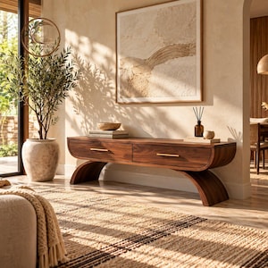 May include: A low-profile wooden media console featuring two drawers with horizontal gold-toned metal pulls, supported by two curved, arch-shaped legs. The piece is crafted from dark wood with a visible grain and has a smooth, rounded rectangular frame.