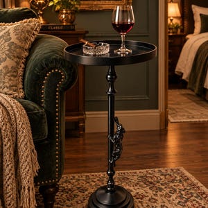 May include: A black metal pedestal side table with a round tray top and a central column featuring a sculpted figure of a climbing animal. The table holds a clear glass wine glass filled with red wine and a glass ashtray with a cigar.