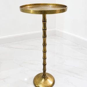 May include: A tall, round side table crafted from brass-toned metal. The table features a flat, circular top with a raised edge, supported by a slender, segmented central column that mimics the appearance of bamboo, resting on a circular base.