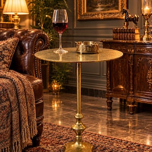 May include: A round gold-toned metal accent table with a textured surface and a pedestal base. The table features a slender central column that flares into a decorative, four-lobed base. The finish has a distressed, metallic gold appearance.