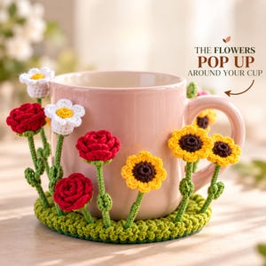 May include: A light pink ceramic mug sits inside a green crocheted base featuring attached three-dimensional flowers including red roses, white daisies, and yellow sunflowers. The text THE FLOWERS POP UP AROUND YOUR CUP is printed above the mug.