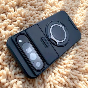May include: A matte black smartphone case featuring a sliding camera lens cover and a circular magnetic ring mount on the back. The case has a rectangular cutout for the camera array and a smooth, solid-colored finish across the entire surface.