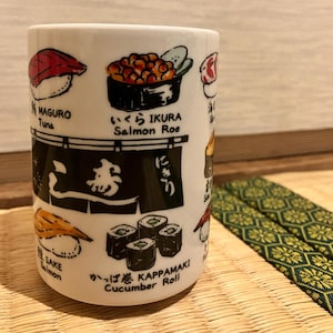 Op de afbeelding: Een witte keramische mok met kleurrijke illustraties van diverse sushisoorten, waaronder Maguro tonijn, Ikura zalmeitjes, Sake zalm en Kappamaki komkommerrolletjes, voorzien van bijbehorende Japanse en Engelse tekstlabels onder elk item.