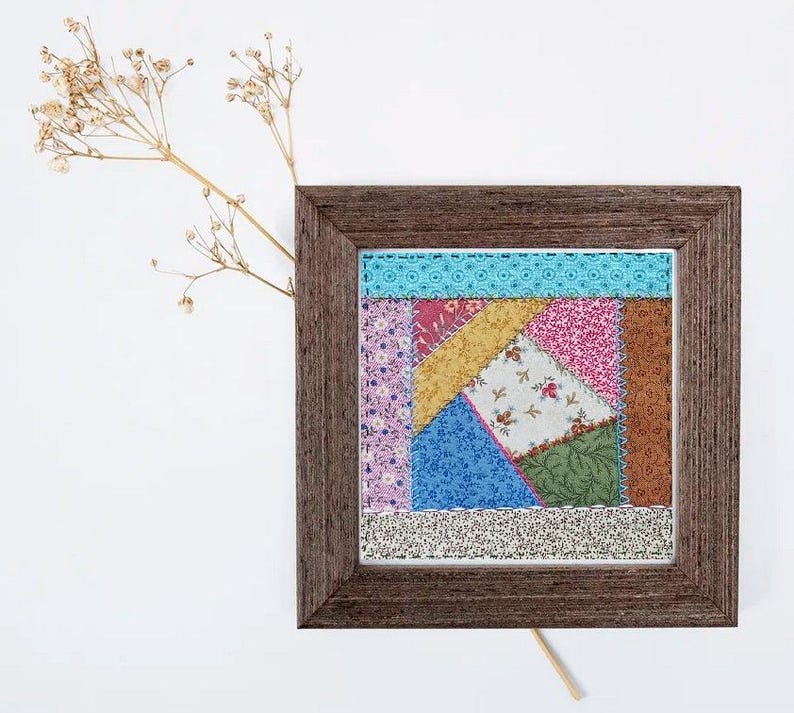 May include: A framed patchwork quilt square with a variety of colorful fabrics, including pink, blue, green, yellow, and brown. The quilt square is surrounded by a brown wooden frame.