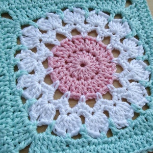 May include: A crocheted square with a pink, white, and light blue design. The center of the square features a pink circle surrounded by white crocheted flowers.