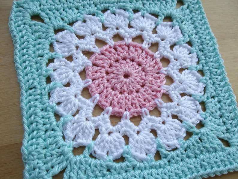 May include: A crocheted square with a pink, white, and light blue design. The center of the square features a pink circle surrounded by white crocheted flowers.