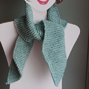 May include: A hand-knitted scarf in a soft, muted teal color. The scarf is displayed on a mannequin, showcasing its triangular ends and textured knit pattern. The scarf appears to be made of a soft yarn.