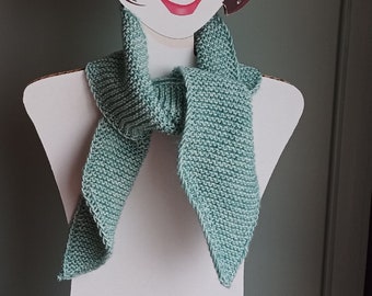 May include: A hand-knitted scarf in a soft, muted teal color. The scarf is displayed on a mannequin, showcasing its triangular ends and textured knit pattern. The scarf appears to be made of a soft yarn.