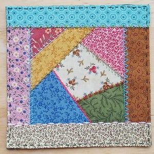 May include: A patchwork quilt block with a variety of floral fabrics in shades of pink, blue, green, and brown. The block is sewn together with a contrasting thread and has a simple, rustic design.