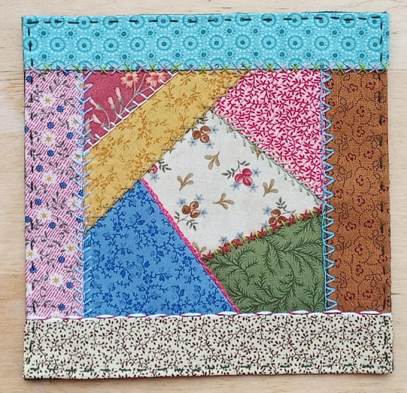 May include: A patchwork quilt block with a variety of floral fabrics in shades of pink, blue, green, and brown. The block is sewn together with a contrasting thread and has a simple, rustic design.