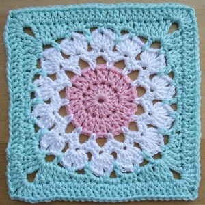 May include: A crocheted square with a pink and white flower design in the center. The square is surrounded by a light blue border.