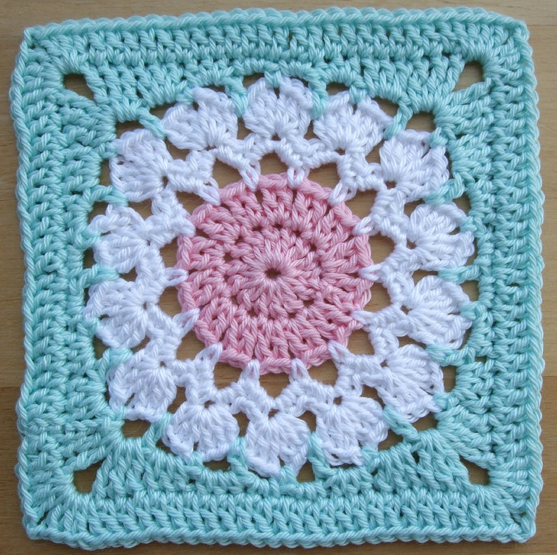 May include: A crocheted square with a pink and white flower design in the center. The square is surrounded by a light blue border.