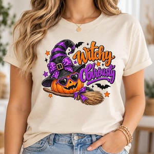 May include: A cream-colored t-shirt featuring a graphic of a jack-o'-lantern wearing a purple and black witch hat and heart-shaped sunglasses. The pumpkin sits on a broomstick with the text "Witchy Obviously" in orange and purple.