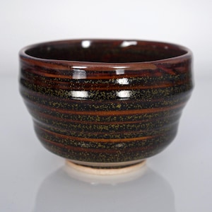 May include: A ceramic bowl with a dark brown and black glaze featuring horizontal ridges and a speckled texture. The bowl has a wide, open mouth and a tapered base that reveals a light beige, unglazed circular foot at the bottom center.