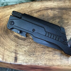 Puede incluir: Cuchillo multiusos negro con una hoja dentada, un abridor de botellas, un cortador de cinturones de seguridad y un rompe cristales. La herramienta tiene un mango negro texturizado y un cuerpo de metal negro.