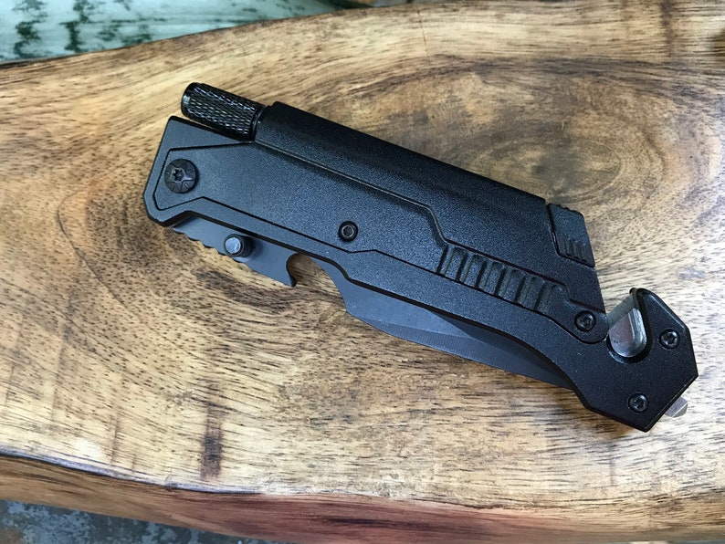 Puede incluir: Cuchillo multiusos negro con una hoja dentada, un abridor de botellas, un cortador de cinturones de seguridad y un rompe cristales. La herramienta tiene un mango negro texturizado y un cuerpo de metal negro.