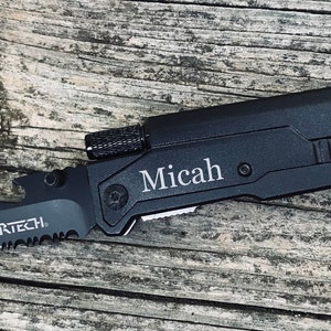Puede incluir: Cuchillo multiusos negro con una hoja dentada, un abridor de botellas, un rompe cristales y un corta cinturones. El cuchillo est&aacute; grabado con el nombre "Micah".