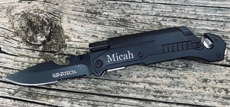 Puede incluir: Cuchillo multiusos negro con una hoja dentada, un abridor de botellas, un rompe cristales y un corta cinturones. El cuchillo est&aacute; grabado con el nombre "Micah".