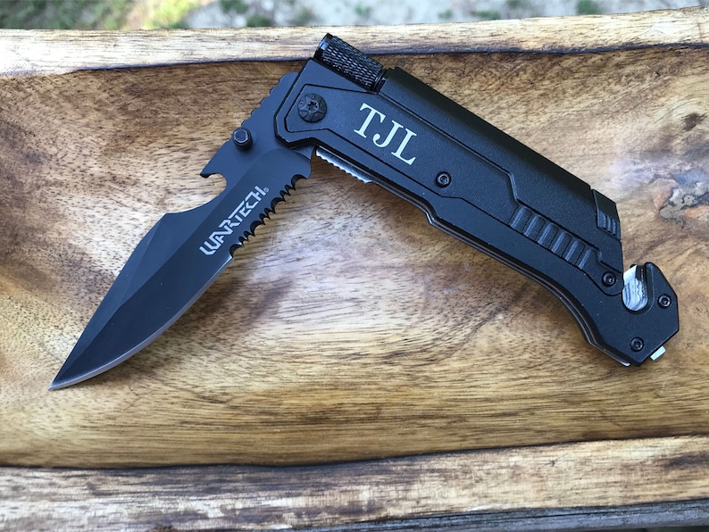 Puede incluir: Un cuchillo de bolsillo multiusos negro con una hoja dentada, abridor de botellas y corta cinturones de seguridad. El cuchillo est&aacute; grabado con las iniciales "TJL".