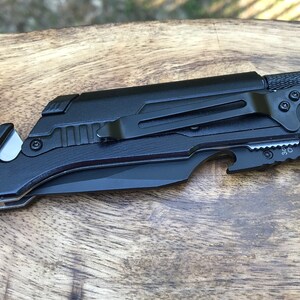 Puede incluir: Cuchillo plegable de bolsillo negro con una cuchilla de seguridad, abridor de botellas y clip para cintur&oacute;n. El cuchillo tiene un mango negro texturizado y una hoja negra.