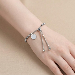 May include: A silver-toned adjustable bracelet featuring a chain of small metallic beads. The bracelet includes a circular charm and two dangling chain ends with spherical beads at the tips, designed with a sliding mechanism for size adjustment.
