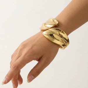 May include: A polished gold-tone cuff bracelet featuring a wide, sculptural design with smooth, organic, curved surfaces. The metal has a high-shine reflective finish that catches the light, creating a bold, solid, and continuous wrap-around form.