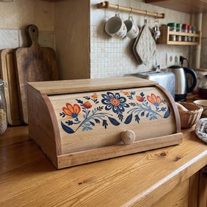 May include: A light-colored wooden bread box with a roll-top lid featuring a painted floral design in shades of blue, orange, and yellow. The box has a small round wooden knob on the front for opening and a smooth, natural wood grain finish.