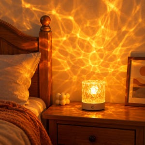 May include: A cylindrical table lamp with a textured, clear glass shade that emits a warm, golden light, creating a rippling water-like projection on the wall. The lamp features a white circular base with a power button icon on the front.