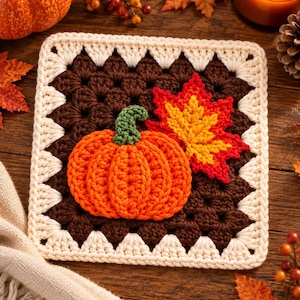 Pode incluir: Um quadrado de crochê com centro castanho escuro, decorado com uma abóbora laranja texturizada e uma folha de ácer vermelha e amarela. O quadrado é contornado por uma borda de crochê em tom creme com um padrão triangular repetitivo.
