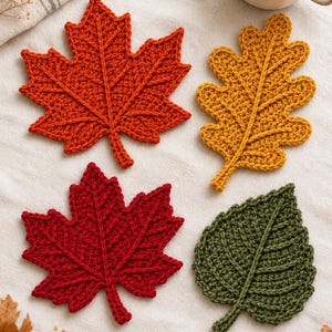 Op de afbeelding: Vier gehaakte bladonderzetters in herfstkleuren. Een oranje esdoornblad, een geel eikenblad, een dieprood esdoornblad en een donkergroen gekarteld blad. Elk item is voorzien van verhoogde centrale nerven en een textuurrijke afwerking.
