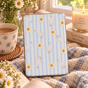 Op de afbeelding: Een rechthoekige tablethoes met een verticaal gestreept patroon in lichtblauw en wit, versierd met herhalende illustraties van witte eenden en gele madeliefjes. De hoes heeft een transparant beschermend frame.