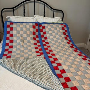 May include: A rectangular quilt featuring a checkerboard pattern of red squares and light gray patterned squares, bordered by a solid blue fabric. The underside of the quilt is visible, showing a light-colored fabric with a small red heart print.