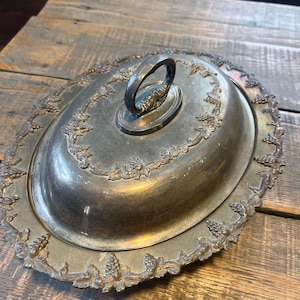 Può includere: Un vassoio da portata ovale in metallo color argento con coperchio a cupola abbinato. Il coperchio presenta una maniglia centrale ad arco e un bordo decorativo in rilievo con grappoli d'uva e foglie di vite. Anche il bordo del vassoio ha il motivo.