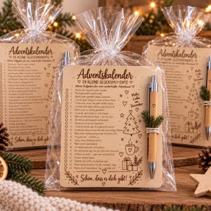 Op de afbeelding: Een houten adventskalenderkaart met 24 genummerde taken, een bamboe pen bevestigd met touw en een klein dennentakje, verpakt in een doorzichtige plastic zak. De kaart bevat gedrukte tekst en feestelijke illustraties.